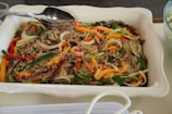 A white rectangular dish filled with a colorful noodle dish, featuring translucent noodles mixed with sliced vegetables such as carrots, bell peppers, and green onions. Mushrooms and sesame seeds are sprinkled on top, with a metal spoon and fork resting on the dish.