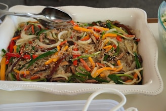 A white rectangular dish filled with a colorful noodle dish, featuring translucent noodles mixed with sliced vegetables such as carrots, bell peppers, and green onions. Mushrooms and sesame seeds are sprinkled on top, with a metal spoon and fork resting on the dish.