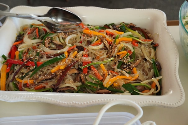 A white rectangular dish filled with a colorful noodle dish, featuring translucent noodles mixed with sliced vegetables such as carrots, bell peppers, and green onions. Mushrooms and sesame seeds are sprinkled on top, with a metal spoon and fork resting on the dish.