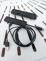 A streaming device with a long USB cable is shown alongside a remote control. Both items are placed on a white surface with a pattern of slots.
