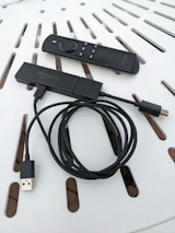 A streaming device with a long USB cable is shown alongside a remote control. Both items are placed on a white surface with a pattern of slots.