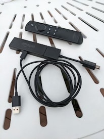 A streaming device with a long USB cable is shown alongside a remote control. Both items are placed on a white surface with a pattern of slots.