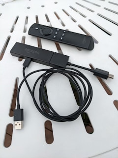 A streaming device with a long USB cable is shown alongside a remote control. Both items are placed on a white surface with a pattern of slots.