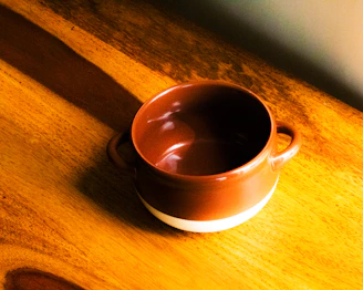 A bright, hand-pulled ceramic mug with warm orange glaze sitting on a wooden table bathed in soft sunlight.