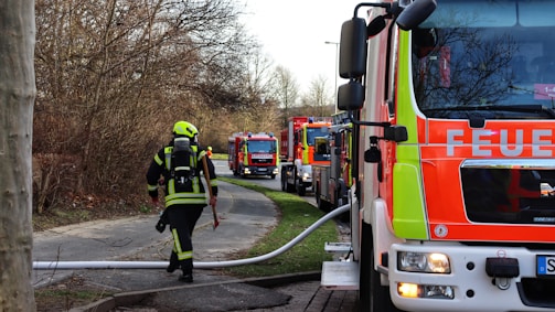 Several fire trucks are parked along a road near a wooded area. A firefighter in protective gear is walking with a hose attached to one of the trucks, surrounded by winter trees and bare branches.