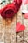 Two red roses are placed on a wooden surface. Near the roses, a small red paper heart is resting on the wood. The heart has small black dots, contributing to a romantic and delicate atmosphere. The wooden background adds a rustic charm to the composition.
