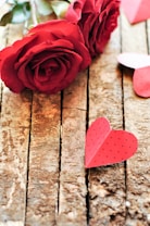 Two red roses are placed on a wooden surface. Near the roses, a small red paper heart is resting on the wood. The heart has small black dots, contributing to a romantic and delicate atmosphere. The wooden background adds a rustic charm to the composition.