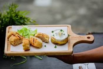 A wooden serving board is presented with grilled bread pieces, fresh green lettuce, and a round container of cheese garnished with parsley. The board is labeled with 'Brasero Steak Frites'. A background of greenery and a blurred area adds to the aesthetic.