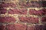 Rough, textured red bricks are stacked in a horizontal pattern, separated by light-colored mortar. The surface of the bricks appears weathered and aged.