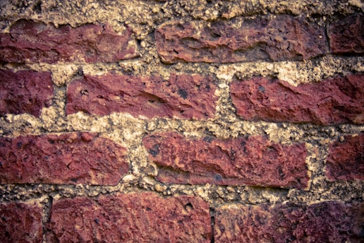 Rough, textured red bricks are stacked in a horizontal pattern, separated by light-colored mortar. The surface of the bricks appears weathered and aged.