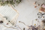 A collection of vintage sheet music is spread out on a textured, light-colored surface, accompanied by dried flowers and leaves. To the right, scissors and spools of thread add a crafting element to the scene.