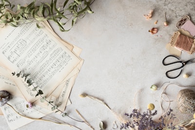 A collection of vintage sheet music is spread out on a textured, light-colored surface, accompanied by dried flowers and leaves. To the right, scissors and spools of thread add a crafting element to the scene.