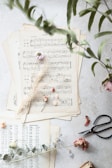 Several sheets of vintage sheet music are arranged on a light surface. Dried flowers, including small roses and delicate leaves, are artistically scattered on top. A pair of black scissors and some green eucalyptus leaves lie near the sheets, adding a touch of nature and craft.