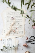 Several sheets of vintage sheet music are arranged on a light surface. Dried flowers, including small roses and delicate leaves, are artistically scattered on top. A pair of black scissors and some green eucalyptus leaves lie near the sheets, adding a touch of nature and craft.