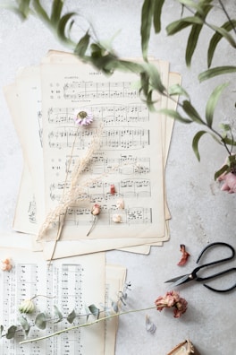 Several sheets of vintage sheet music are arranged on a light surface. Dried flowers, including small roses and delicate leaves, are artistically scattered on top. A pair of black scissors and some green eucalyptus leaves lie near the sheets, adding a touch of nature and craft.