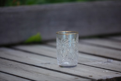 A clear glass with ornate etched patterns is placed on a wooden surface. It features a gold rim and intricate leaf-like designs on its body. The background is slightly blurred, giving an impression of an outdoor setting.