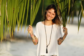 A woman with tattoos on her arms stands outdoors, holding large palm leaves on either side of her face. She is smiling and wearing a white shirt and a long black beaded necklace. The background features blurred greenery and sandy terrain, creating a tropical and relaxed ambiance.