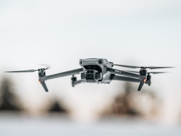 A sleek, modern drone captured in mid-flight against a blurred background. Its design features four propellers and a camera mounted at the front, embodying a high-tech aesthetic.