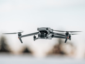 A sleek, modern drone captured in mid-flight against a blurred background. Its design features four propellers and a camera mounted at the front, embodying a high-tech aesthetic.