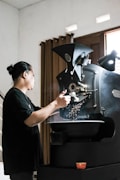 A person is operating a large coffee roasting machine, with beans visible inside the machine's hopper. The room is softly lit, with curtains in the background and a warm, ambient feel. There is a small red bowl on a surface in the foreground.