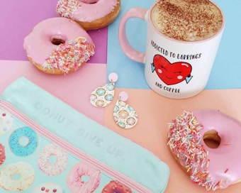 A pastel-themed flat lay with three pink frosted donuts covered in colorful sprinkles, a mug with a cartoon heart graphic labeled 'addicted to earrings and coffee', tear-shaped earrings with donut patterns, and a pouch with a donut design and the phrase 'DONUT GIVE UP!'