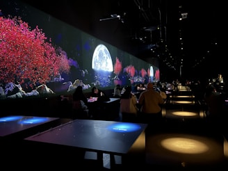 The image features a dimly lit indoor setting with long tables and people seated. A vibrant digital display on the wall shows a serene scene with red-leaved trees, rocky terrain, and a full moon over water, suggesting an immersive experience.