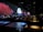 The image features a dimly lit indoor setting with long tables and people seated. A vibrant digital display on the wall shows a serene scene with red-leaved trees, rocky terrain, and a full moon over water, suggesting an immersive experience.