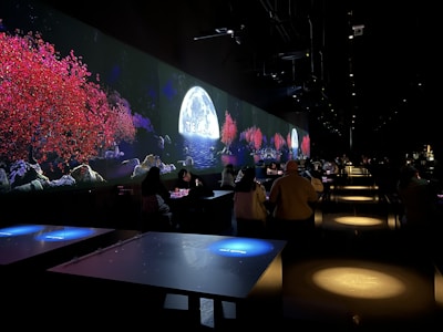 The image features a dimly lit indoor setting with long tables and people seated. A vibrant digital display on the wall shows a serene scene with red-leaved trees, rocky terrain, and a full moon over water, suggesting an immersive experience.