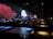 The image features a dimly lit indoor setting with long tables and people seated. A vibrant digital display on the wall shows a serene scene with red-leaved trees, rocky terrain, and a full moon over water, suggesting an immersive experience.