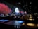 The image features a dimly lit indoor setting with long tables and people seated. A vibrant digital display on the wall shows a serene scene with red-leaved trees, rocky terrain, and a full moon over water, suggesting an immersive experience.