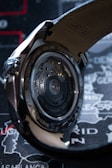 A close-up view of a wristwatch showcasing the intricate mechanics visible through its transparent case back. The metallic components gleam under the light, with engraved text on the edge detailing specifications. The background features a map design with faint geographical labels.