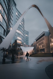 An urban scene with modern architecture featuring sleek, glass buildings and a prominent sculptural arch. A person walks along the curved sidewalk, with small palm trees in pots adding greenery to the surroundings.