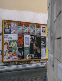 A wooden display board on a street corner covered with a variety of colorful posters, advertisements, and event announcements. The posters feature artistic graphics, text about exhibitions and performances, and different cultural events. The board is situated against a worn, slightly graffiti-marked wall, and the scene is part of an urban setting with cobblestone pavement.