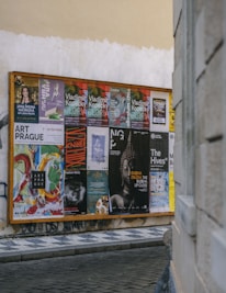 A wooden display board on a street corner covered with a variety of colorful posters, advertisements, and event announcements. The posters feature artistic graphics, text about exhibitions and performances, and different cultural events. The board is situated against a worn, slightly graffiti-marked wall, and the scene is part of an urban setting with cobblestone pavement.