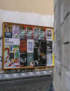 A wooden display board on a street corner covered with a variety of colorful posters, advertisements, and event announcements. The posters feature artistic graphics, text about exhibitions and performances, and different cultural events. The board is situated against a worn, slightly graffiti-marked wall, and the scene is part of an urban setting with cobblestone pavement.