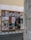 A wooden display board on a street corner covered with a variety of colorful posters, advertisements, and event announcements. The posters feature artistic graphics, text about exhibitions and performances, and different cultural events. The board is situated against a worn, slightly graffiti-marked wall, and the scene is part of an urban setting with cobblestone pavement.