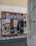 A wooden display board on a street corner covered with a variety of colorful posters, advertisements, and event announcements. The posters feature artistic graphics, text about exhibitions and performances, and different cultural events. The board is situated against a worn, slightly graffiti-marked wall, and the scene is part of an urban setting with cobblestone pavement.