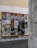 A wooden display board on a street corner covered with a variety of colorful posters, advertisements, and event announcements. The posters feature artistic graphics, text about exhibitions and performances, and different cultural events. The board is situated against a worn, slightly graffiti-marked wall, and the scene is part of an urban setting with cobblestone pavement.