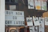 A storefront window display features a large sign with bold letters that reads 'Buy Now or Cry Later'. Above the sign, there are small cards clipped with clothespins, displaying illustrations and artwork, including a donkey, a sun, and various landscapes. The scene includes some merchandise, hinting at a retail environment.