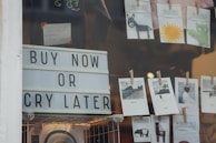 A storefront window display features a large sign with bold letters that reads 'Buy Now or Cry Later'. Above the sign, there are small cards clipped with clothespins, displaying illustrations and artwork, including a donkey, a sun, and various landscapes. The scene includes some merchandise, hinting at a retail environment.