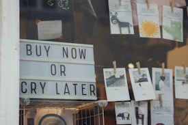 A storefront window display features a large sign with bold letters that reads 'Buy Now or Cry Later'. Above the sign, there are small cards clipped with clothespins, displaying illustrations and artwork, including a donkey, a sun, and various landscapes. The scene includes some merchandise, hinting at a retail environment.