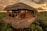 A rustic, thatched-roof treehouse is nestled among tropical greenery at sunset. It has a wooden structure with open sides, giving it a natural and harmonious appearance. A person is meditating on the balcony, adding a serene and peaceful element to the scene. Another person is seen standing on a platform, observing the surroundings.
