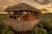 A rustic, thatched-roof treehouse is nestled among tropical greenery at sunset. It has a wooden structure with open sides, giving it a natural and harmonious appearance. A person is meditating on the balcony, adding a serene and peaceful element to the scene. Another person is seen standing on a platform, observing the surroundings.