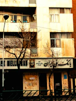 A building facade with a bank on the ground floor, featuring signs in a non-Latin script. Bare trees are in front of the building, indicating a winter or late autumn setting. On the balcony above, several potted plants can be seen.