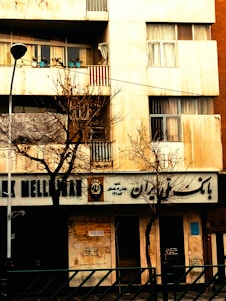 A building facade with a bank on the ground floor, featuring signs in a non-Latin script. Bare trees are in front of the building, indicating a winter or late autumn setting. On the balcony above, several potted plants can be seen.