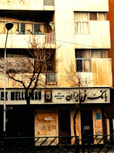 A building facade with a bank on the ground floor, featuring signs in a non-Latin script. Bare trees are in front of the building, indicating a winter or late autumn setting. On the balcony above, several potted plants can be seen.