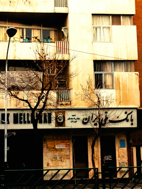 A building facade with a bank on the ground floor, featuring signs in a non-Latin script. Bare trees are in front of the building, indicating a winter or late autumn setting. On the balcony above, several potted plants can be seen.