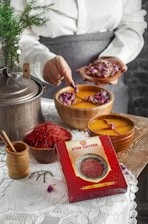 A person is holding a small plate of dried rosebuds, using a spoon to add them to a bowl of liquid with floating roses. A box labeled 'Atich Saffron' is prominently placed in front. On a wooden surface, there are various saffron containers, a metal pot, and a wooden grinder. A white lace tablecloth and a small evergreen branch add to the rustic setting.