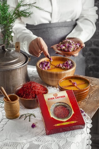 A person is holding a small plate of dried rosebuds, using a spoon to add them to a bowl of liquid with floating roses. A box labeled 'Atich Saffron' is prominently placed in front. On a wooden surface, there are various saffron containers, a metal pot, and a wooden grinder. A white lace tablecloth and a small evergreen branch add to the rustic setting.