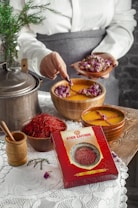 A person is holding a small plate of dried rosebuds, using a spoon to add them to a bowl of liquid with floating roses. A box labeled 'Atich Saffron' is prominently placed in front. On a wooden surface, there are various saffron containers, a metal pot, and a wooden grinder. A white lace tablecloth and a small evergreen branch add to the rustic setting.
